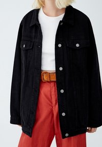 Schwarze Denimjacke mit klassischem Schnitt, ausgestattet mit zwei Fronttaschen, silbernen Knöpfen und entspannter Passform, getragen über einem weißen Oberteil und roten Hosen.