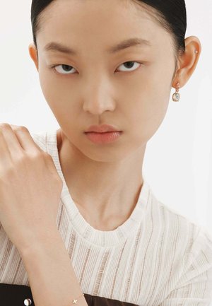 Pendientes de cristal transparente con un cierre de plata, usados por un modelo con una blusa blanca texturizada que presenta rayas horizontales y un cuello redondo.