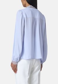 Blusa a maniche lunghe blu chiaro con dettagli in pizzo sul colletto e sui polsini. Vestibilità ampia con pieghe sul retro. Tessuto morbido e liscio.