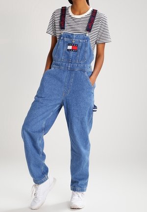 Osoba ubrana w niebieskie ogrodniczki dżinsowe z logo Tommy Jeans, czarno-białą pasiastą koszulkę i białe trampki, stojąca z rękami w kieszeniach.