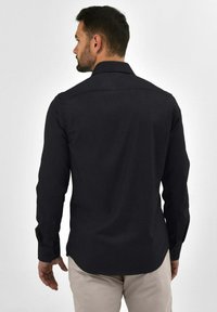 Zwarte langemouwen shirt met een standaard kraag, knoopmanchetten en een gladde textuur, gezien vanuit de achterkant tegen een lichte achtergrond.