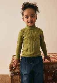 Grüner Kabelstrick-Turtleneck-Pullover mit langen Ärmeln, kombiniert mit dunklen Jeans. Getragen von einem lächelnden Kind, dessen Hände auf einer Holztruhe ruhen.
