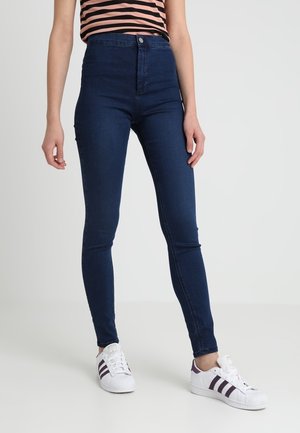 Femme portant un jean skinny bleu marine taille haute, un haut rayé rose et noir, et des baskets blanches avec des rayures noires.