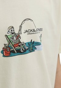 T-shirt en coton beige clair avec un graphique représentant un squelette en train de pêcher depuis une chaise, avec un seau et des éléments aquatiques dans le design.