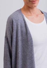 Grijze cardigan gemaakt van zachte, gebreide stof. Heeft een open voorkant en een relaxte pasvorm. Draagt over een wit T-shirt met een ronde hals.