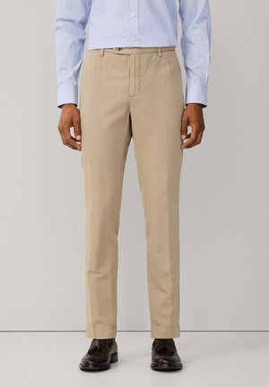 Hombre con pantalones beige de pierna recta, camisa azul claro con botones y mocasines marrón oscuro con borlas, de pie contra un fondo liso.