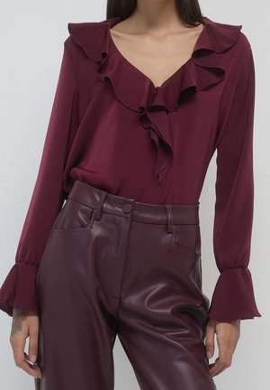 Femme portant un chemisier bordeaux à manches longues avec un col et des poignets volantés, associé à un pantalon en cuir bordeaux taille haute.