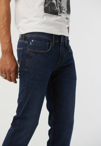 Jeans en denim bleu foncé avec une coupe slim, présentant des poches classiques et des détails de couture subtils. Portés avec un T-shirt blanc imprimé.