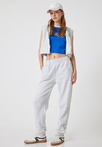 Grijze sweatpants, blauwe crop top, en een grijze mouwloze hoodie, gecombineerd met een witte pet en witte sneakers met grijze en zwarte accenten.