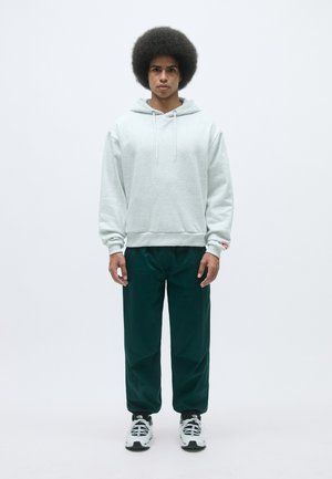 Világosszürke kapucnis pulóver kenguru zsebbel, húzózsinórral és bordázott mandzsettákkal, sötétzöld nadrággal és fehér sportcipővel párosítva.
