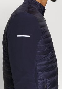 Mörkblå puffer-väst med en slät, quiltad textur; har en hög krage, en ficka med dragkedja på sidan och en reflexrand på ärmen.