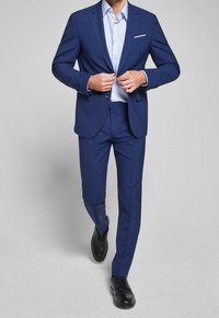 Completo blu con pantaloni abbinati, vestibilità su misura, giacca monopetto, camicia azzurra sotto, completato da scarpe eleganti nere.