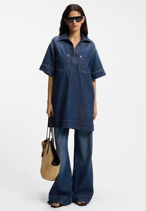 Moteris dėvi per didelę džinsinę palaidinę, plačias džinsines kelnes, juodus sandalus, saulės akinius, laiko smėlio spalvos pintinę rankinę su juodomis rankenomis.