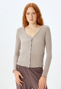 Ribbet cardigan i lys taupe med V-hals, knapfront og bølgede kanter på ærmer og kant, lavet af et blødt materiale.