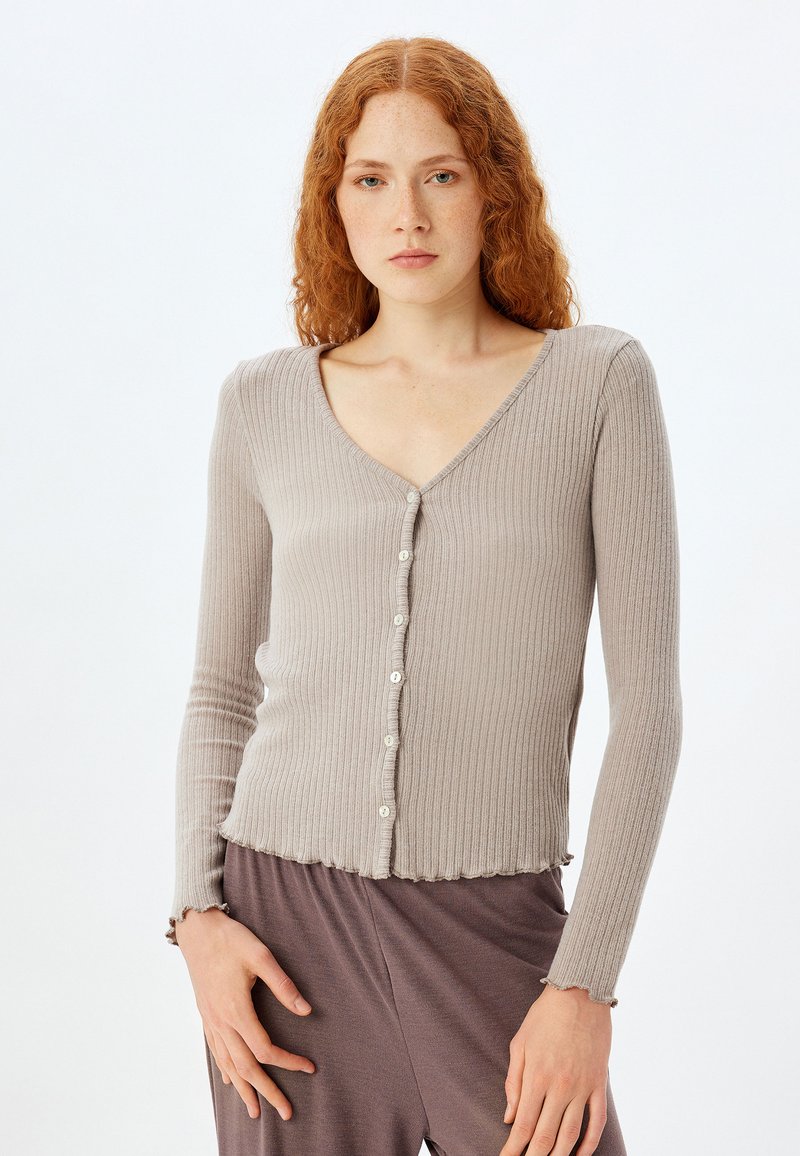 Ribbet cardigan i lys taupe med V-hals, knapfront og bølgede kanter på ærmer og kant, lavet af et blødt materiale.