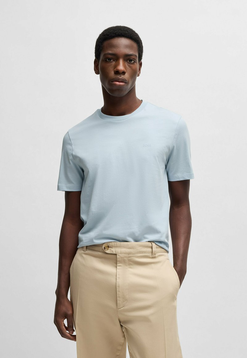Light blue cotton t-shirt with a crew neckline, short sleeves, and subtle logo detail, paired with beige trousers featuring a button closure.
