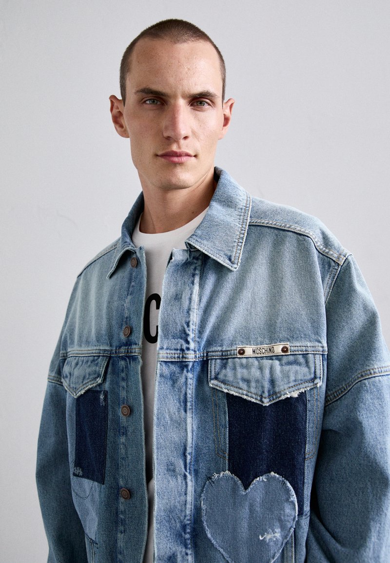 Jonge man met kort haar, gekleed in een lichtblauw denimjack met donkere vlekken en een hartvormige patch op de voorkant.