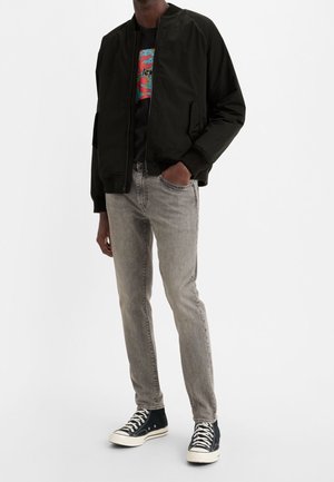 Person trägt eine schwarze Bomberjacke, ein schwarzes T-Shirt mit Grafik, graue schlanke Jeans und schwarze High-Top-Sneaker mit weißen Schnürsenkeln vor weißem Hintergrund.