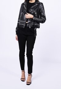 Veste courte de motard en cuir noir avec détails cloutés, fermetures éclair argentées et poches latérales. Associée à un pantalon noir slim et des talons noirs à brides.