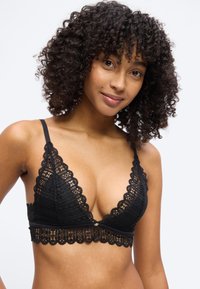 Γυναίκα με σγουρά μαλλιά που φοράει μαύρο δαντελένιο bralette, χαμογελώντας απαλά μπροστά σε ένα απλό ανοιχτό φόντο.