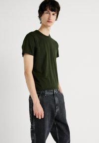Joven con una camiseta de manga corta verde olivo y unos pantalones vaqueros negros holgados, de pie contra un fondo blanco liso.