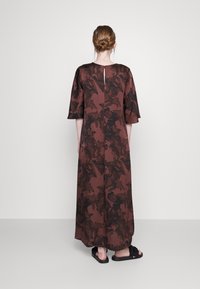 Vestido longo castanho com um padrão abstrato texturizado, mangas curtas e uma abertura em forma de chave nas costas. Usado com sandálias pretas.