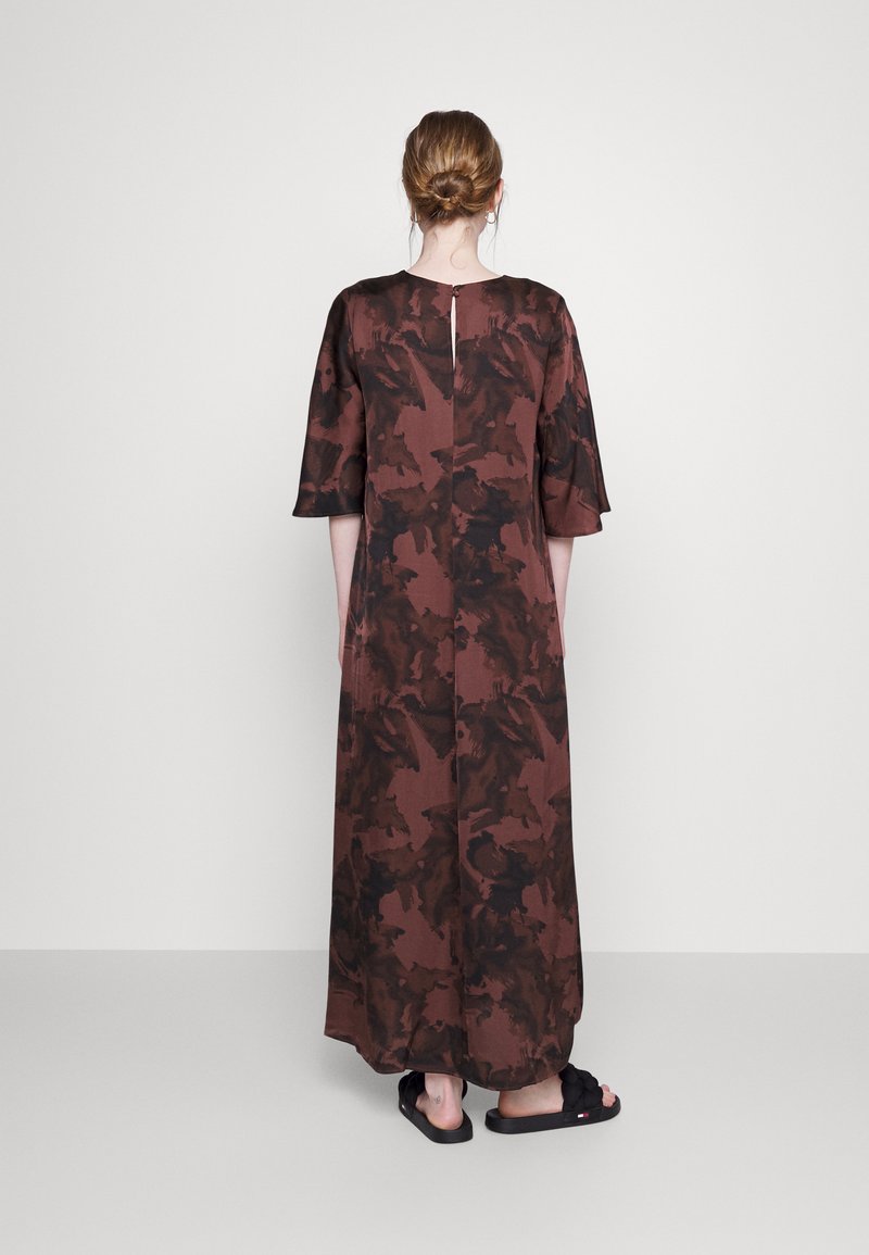 Vestido longo castanho com um padrão abstrato texturizado, mangas curtas e uma abertura em forma de chave nas costas. Usado com sandálias pretas.