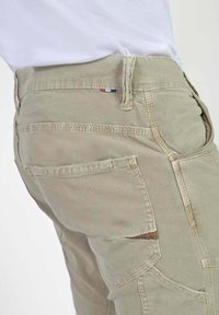 Beige katoenen cargobroek met meerdere zakken. Bevat zichtbare stiksels en een klein driekleurig vlagaccent bij de taille. De textuur lijkt stevig.