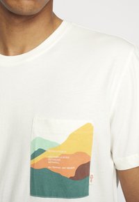 Witte t-shirt met korte mouwen en een kleurrijk zakontwerp met golven in groen, blauw, oranje en geel. Zachte stoftextuur.