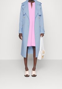 Niebieski płaszcz woskowy z podwójnymi guzikami na różowej sukience. Beżowe klapki z odkrytą palcą. Biała torebka z koralikami i słomkowymi akcentami.