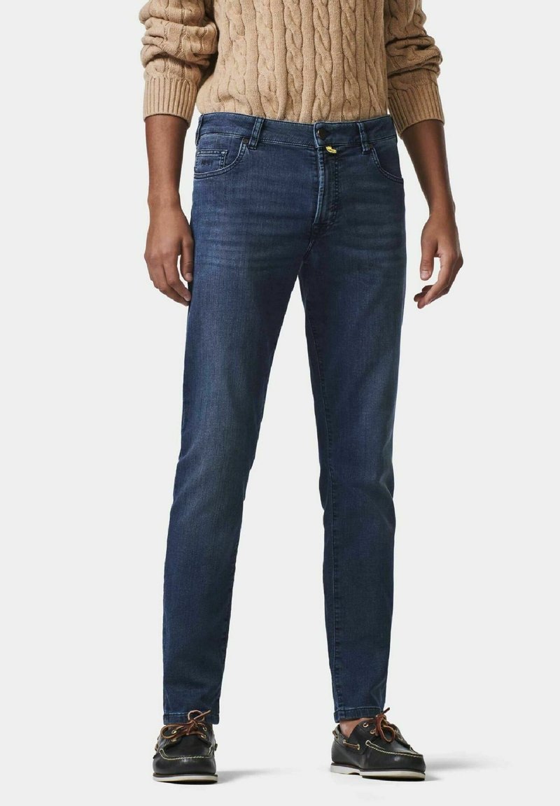 Jean en denim bleu foncé avec une coupe droite, un design à cinq poches et des détails en métal jaune. Porté avec un pull en laine torsadée beige et des chaussures noires.