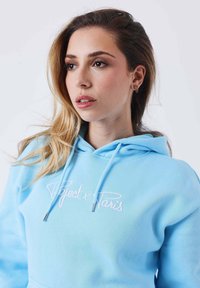 Felpa blu chiaro con cappuccio e cordino, con la scritta ricamata "Project x Paris" in rosa. Presenta una texture morbida e un design a vestibilità rilassata.