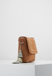 Tan nahkalaukku, jossa on läppäkilpi ja koristeellinen vihreä ja kerma tasseli. Kompakti, suorakulmainen muoto, jossa on näkyvät ompeleet.