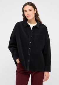 Camisa de pana negra con cuello tipo solapa, bolsillo frontal y cierre de botones. Presenta un corte relajado y textura ribeteada.