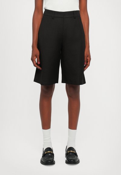 Schwarze maßgeschneiderte Shorts mit geradem Schnitt, aus leichtem Stoff gefertigt. Mit einer mittelhohen Taille und scharfen Kanten. Kombiniert mit schwarzen Loafern.