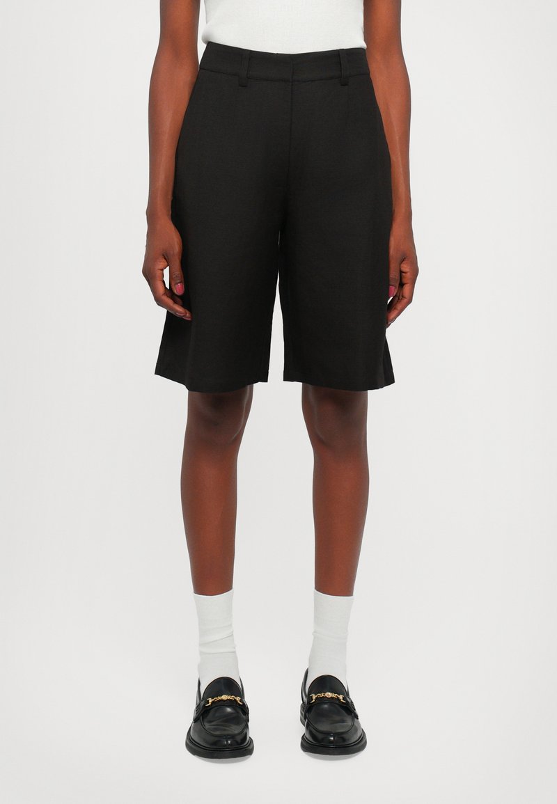 Sorte skreddersydde shorts med rett passform, laget av lett materiale. Har midjehøyde og skarpe kanter. Kombinert med sorte loafers.