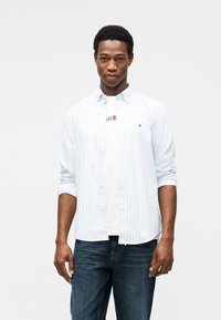 Πουκάμισο με μπλε ανοιχτό και άσπρες ρίγες και κουμπιά πάνω από ένα λευκό t-shirt, συνδυασμένο με μπλε τζιν. Διαθέτει ένα μικρό λογότυπο στο πουκάμισο.