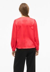 Blusa de satén roja con mangas largas abullonadas, escote redondo y puños fruncidos, combinada con pantalones negros. Textura suave y color vibrante.