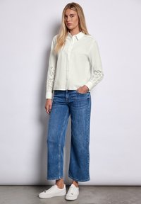 Camisa blanca de manga larga con detalles perforados en las mangas, combinada con jeans de mezclilla azul de pierna ancha y zapatillas blancas, sobre un fondo neutro.