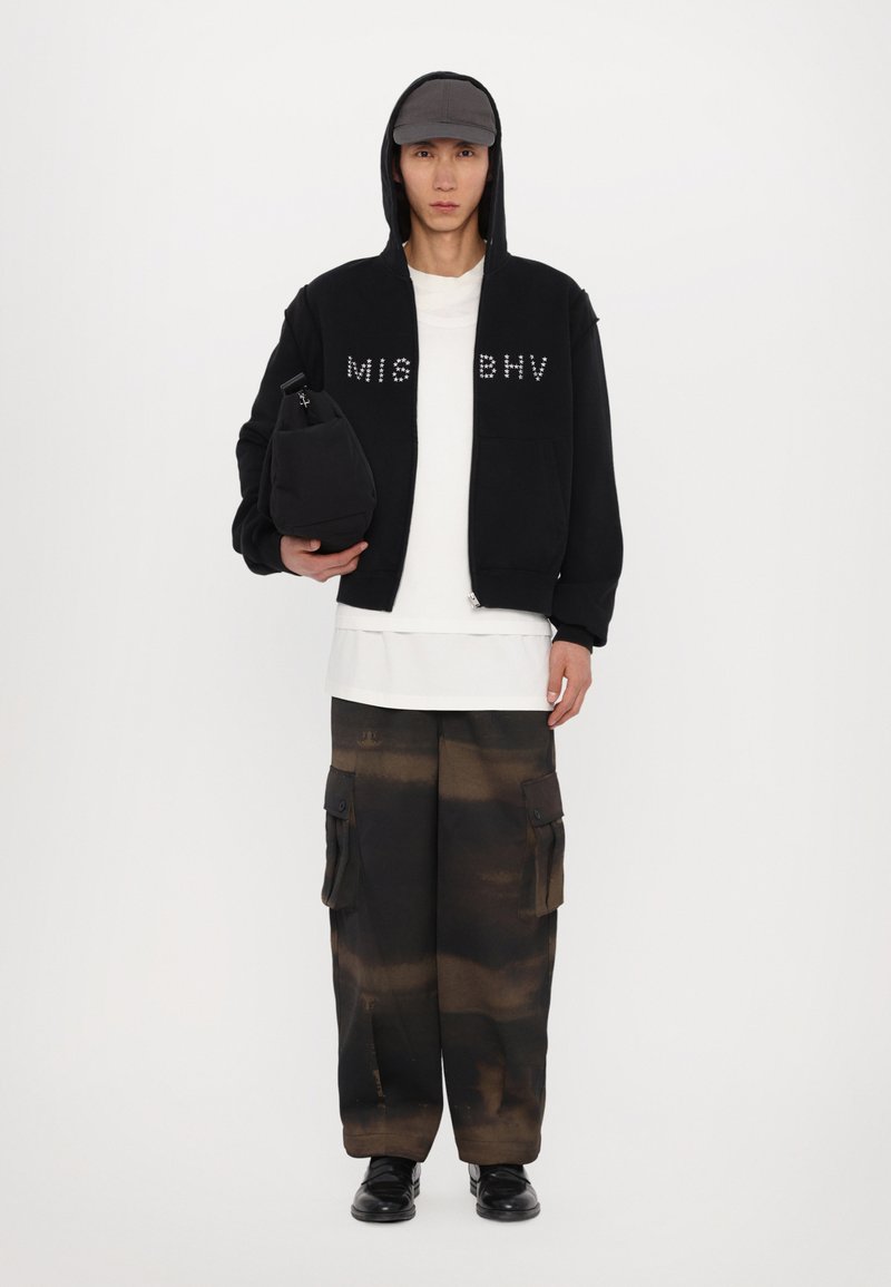 Black hoodie with studded lettering, layered over a white t-shirt, paired with oversized brown tie-dye cargo pants and black shoes.