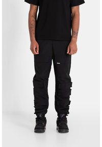 Černé cargo kalhoty s mnoha kapsami, texturovaná tkanina, volný střih a vrstvené detaily. V kombinaci s černými teniskami s síťovými prvky.