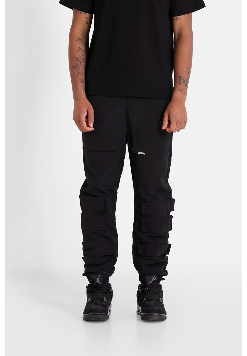 Černé cargo kalhoty s mnoha kapsami, texturovaná tkanina, volný střih a vrstvené detaily. V kombinaci s černými teniskami s síťovými prvky.