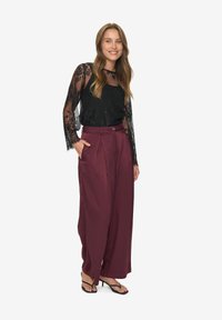 Woman smiling wearing a sheer black lace long-sleeve top, high-waisted wide-leg burgundy pants, and black open-toe heeled sandals.