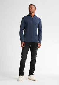 Top lungo a maniche lunghe con zip di colore blu navy, caratterizzato da un motivo testurizzato, abbinato a jeans neri e sneakers bianche. Il modello è in piedi su uno sfondo chiaro.