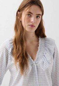 Giovane donna con lunghi capelli castani che indossa una camicia a scacchi bianca e blu con colletto arricciato, che guarda direttamente nella telecamera.
