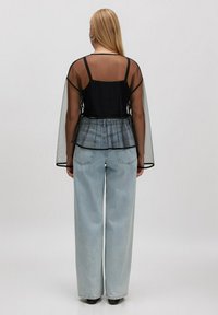Frau mit langen blonden Haaren, die ein schwarzes transparentes Oberteil über einem schwarzen Tanktop und hellblauen weit geschnittenen Jeans trägt, steht vor einem schlichten Hintergrund.