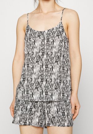 Frau trägt ein ärmelloses Oberteil mit schwarz-weißem Muster und passende Shorts mit dünnen Schulterriemen, steht vor schlichter Hintergrund.