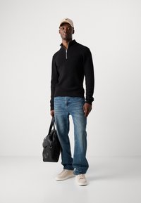 Black zip-up sweater with ribbed texture, light blue jeans, cream sneakers, and a beige cap with a logo; holding a black bag.