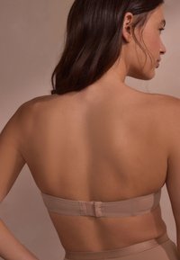 Donna con capelli lunghi e scuri che indossa un reggiseno beige senza spalline, mostrata di spalle su uno sfondo neutro.