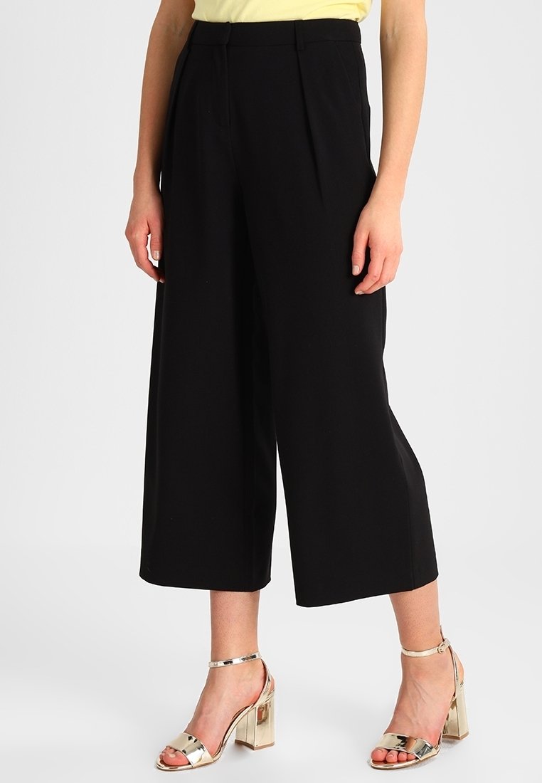 Femme portant un pantalon noir large et court et des sandales dorées à talons hauts, debout avec les mains détendues le long du corps.